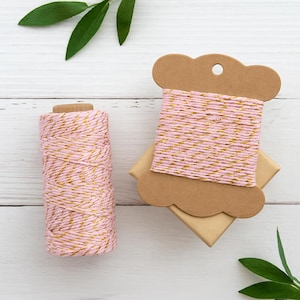 May include: A spool of pink and gold metallic twine alongside a cylindrical roll of the same pink and gold twine. The twine features a twisted texture with alternating strands of soft pink cotton and shimmering gold metallic thread throughout.