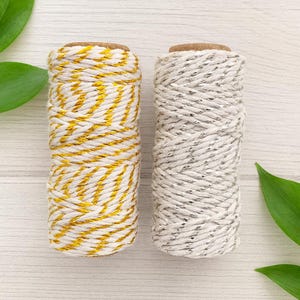 May include: Two cylindrical spools of twine. The left spool features white cord twisted with metallic gold thread. The right spool features white cord twisted with metallic silver thread. Both spools have a brown cardboard core at the top.