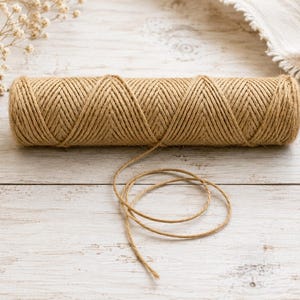 May include: A cylindrical spool of natural tan jute twine featuring a distinct diamond-shaped pattern created by the winding of the cord. A single loose strand of the brown fibre extends from the centre of the spool, curling onto the surface.