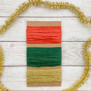 May include: Three strips of crinkled paper ribbon wrapped around a rectangular piece of brown cardboard. The top ribbon is bright red, the middle ribbon is dark forest green, and the bottom ribbon is a metallic gold colour.