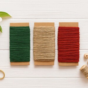 May include: Three rectangular cardboard spools wrapped with dark green, natural tan, and deep red twine. Included are gold-toned metal scissors, a small wooden spool of tan twine, and six small, round, light-coloured wooden beads for crafting.