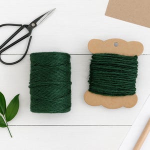 May include: A pair of black metal scissors, a cylindrical spool of dark green twine, a flat cardboard bobbin wrapped in dark green twine, a small green leaf sprig, a wooden pencil, and a piece of brown paper on a white wooden surface.