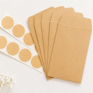 Puede incluir: Un conjunto de cinco sobres rectangulares de papel kraft de color marr&oacute;n claro dispuestos en forma de abanico, acompa&ntilde;ados por dos tiras de etiquetas adhesivas circulares del mismo color y una peque&ntilde;a rama de flores blancas.