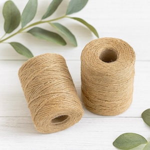 May include: Two cylindrical spools of natural tan jute twine. Each spool features a tightly wound, textured brown fibre cord wrapped around a central cardboard core. The twine has a rustic, fibrous appearance with visible strands throughout the surface.