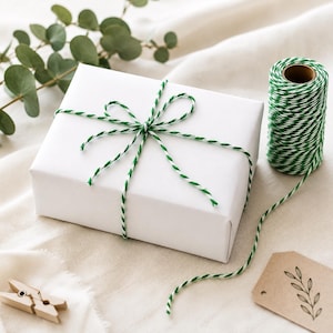 May include: A rectangular gift box wrapped in plain white paper, tied with green and white baker's twine in a bow. Beside the box sits a spool of the same green and white twine, a small wooden clothespin, and a tan gift tag with a leaf illustration.