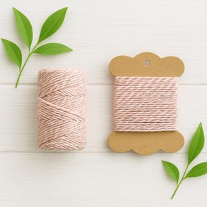 May include: A spool of light pink and white twisted cotton twine alongside a matching ball of twine. The twine is wound tightly in a multi-ply construction, featuring a soft pastel colour palette. Two small green leafy sprigs are placed nearby.