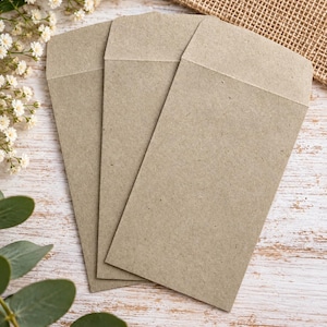 Może przedstawiać: Trzy prostokątne koperty wykonane z jasnobrązowego, teksturowanego papieru z recyklingu. Każda koperta ma spiczastą klapkę u g&oacute;ry i pionową orientację. Papier ma wł&oacute;knisty, nakrapiany wygląd charakterystyczny dla naturalnego materiału typu kraft.