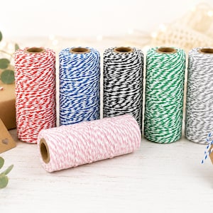 May include: Six cylindrical spools of baker's twine in red, blue, black, green, grey, and pink, each featuring a white twisted pattern. The spools are arranged in a row with one spool lying horizontally in front, accompanied by brown gift tags.