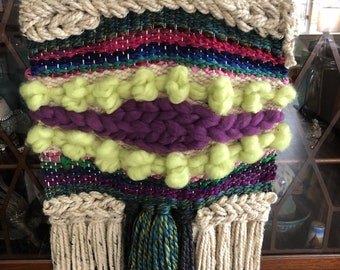 May include: A woven wall hanging with a colorful striped pattern, a purple and green section with fluffy yarn balls, and a fringe border. The hanging is attached to a wooden dowel.