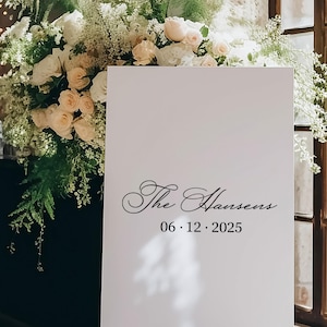 以下が含まれることがあります： 木製のイーゼルに立てられた白い看板。黒い筆記体で「The Hansens」と日付06.12.2025と書かれています。白いバラとピーチ色のバラ、緑の葉で飾られた大きなフラワーアレンジメントが特徴です。