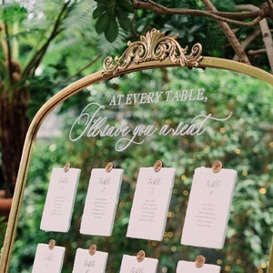 May include: A gold-framed mirror with a white inscription that reads "At every table, I'll save you a seat". The mirror is displaying a seating chart with eight cards, each labelled with a table number.