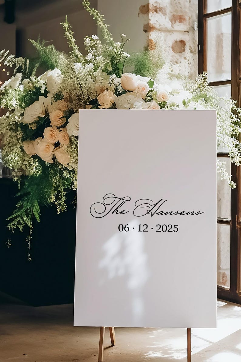 以下が含まれることがあります： 木製のイーゼルに立てられた白い看板。黒い筆記体で「The Hansens」と日付06.12.2025と書かれています。白いバラとピーチ色のバラ、緑の葉で飾られた大きなフラワーアレンジメントが特徴です。
