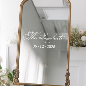 May include: A gold-framed mirror with an ornate design, standing on a black easel. The mirror has a white inscription that reads "The Lamberts 06.12.2025".