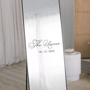 May include: A full-length mirror with a black frame, standing on a black stand. The mirror has the text "The Ugaras" and the date "06.12.2025" in a decorative font. The background includes a white curtain and a small white table.