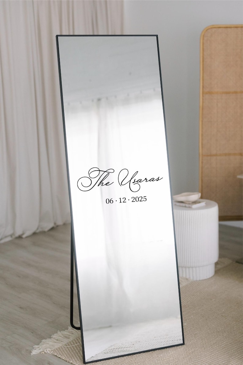 May include: A full-length mirror with a black frame, standing on a black stand. The mirror has the text "The Ugaras" and the date "06.12.2025" in a decorative font. The background includes a white curtain and a small white table.