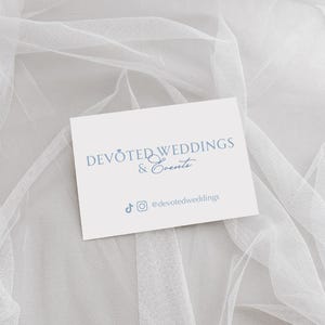 以下が含まれることがあります： 白地の名刺に、ソフトブルーのフォントで「DEVOTED WEDDINGS & Events」と書かれています。カードには、TikTokとInstagramのソーシャルメディアアイコンと、ハンドル名@devotedweddingsも含まれています。カードは白く透き通った生地の上に置かれています。