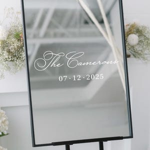 May include: A rectangular mirror with a black frame, mounted on a black easel. The mirror reflects a bright interior. White script reads "The Camerons" above the date "07.12.2025". Decorative floral arrangements flank the mirror.