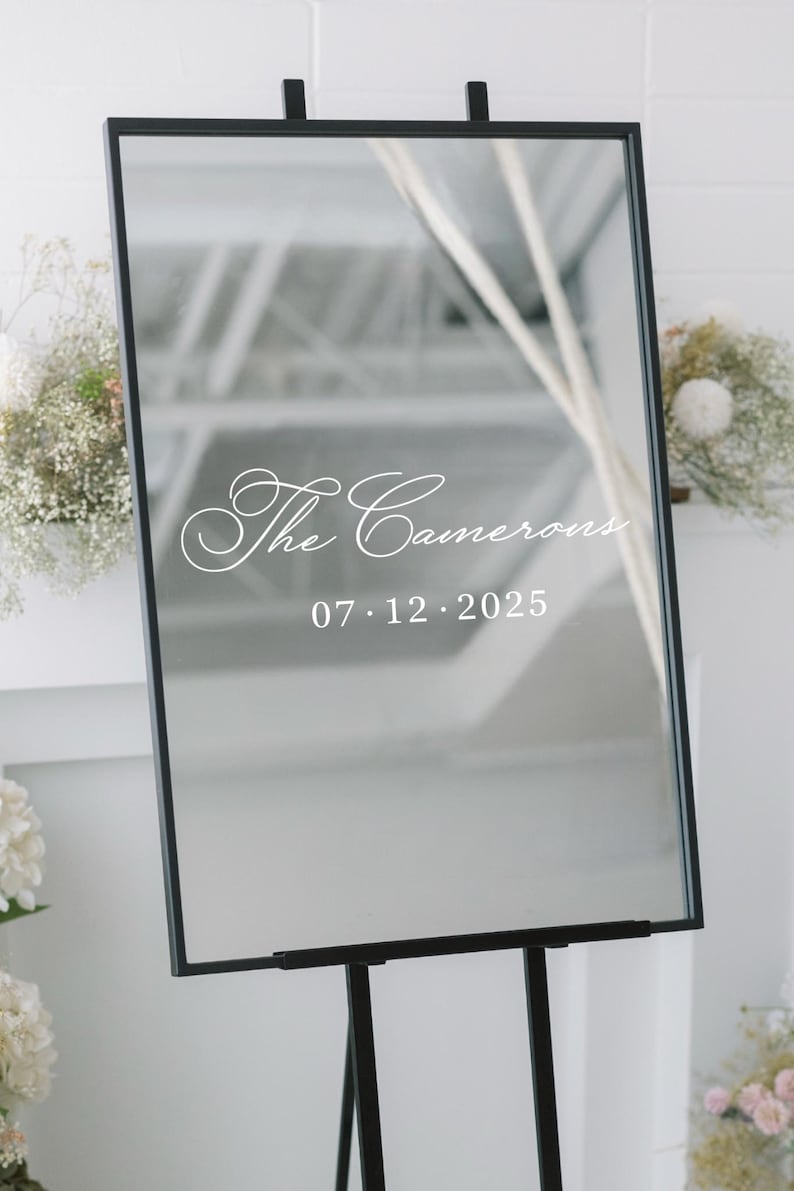 May include: A rectangular mirror with a black frame, mounted on a black easel. The mirror reflects a bright interior. White script reads "The Camerons" above the date "07.12.2025". Decorative floral arrangements flank the mirror.