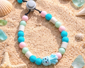 May include: A beaded bracelet with a beach theme. The bracelet features turquoise, pink, white, and mint green beads, and a blue jellyfish charm. The bracelet is displayed on sand with seashells and a starfish. The bracelet is adjustable.