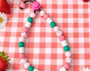 May include: A necklace with pink, green, and white beads, set against a red and white gingham background. The necklace has a pink clasp. Strawberries and daisies are arranged around the necklace.