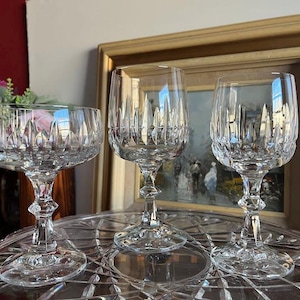 May include: Three clear crystal wine glasses with intricate cut patterns. The glasses are on a decorative glass tray, with a framed painting in the background. The glasses have a classic, elegant design.