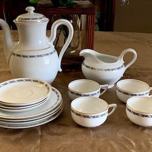 Puede incluir: Un juego de té de porcelana blanca con ribete dorado y un patrón floral gris. El juego incluye una tetera, una jarra de crema y seis tazas y platillos.