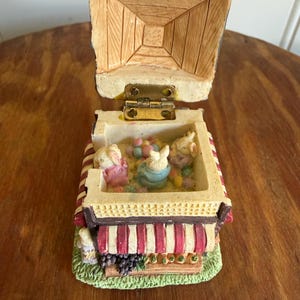 May include: A decorative music box with a hinged lid, depicting a miniature scene of three bunnies inside a shop. The shop has a red and white striped awning and a green base with grapes and vegetables. The box is open, revealing the interior.