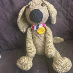 Op de afbeelding: Een gehaakt knuffeldier in de vorm van een hond met een bruin lichaam, een zwarte neus en bruine ogen. De hond draagt een kleurrijk gehaakt halsband met een gele bloem.