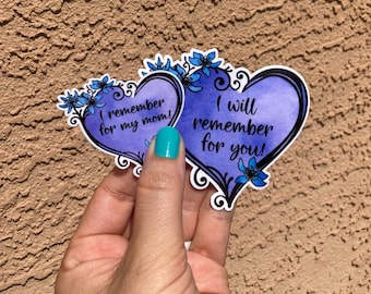 May include: Two heart-shaped stickers with watercolor-style purple backgrounds and black outlines. The stickers are decorated with blue floral accents and the phrases "I remember for my mom!" and "I will remember for you!" are written on them.