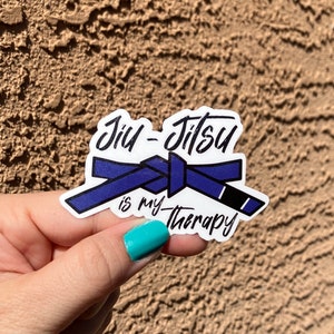 Op de afbeelding: Een witte sticker met een blauw en zwart Jiu-Jitsu-gordelontwerp. De tekst "Jiu-Jitsu is my therapy" is in zwart geschreven.