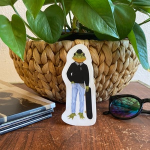 May include: A sticker featuring a frog dressed in a black sweater, plaid shirt, blue jeans, and a bowler hat, holding a skateboard. The frog has a green face and webbed feet. The sticker is on a wooden surface.