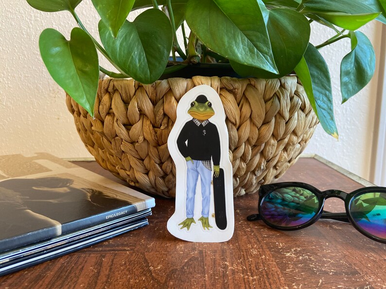 May include: A sticker featuring a frog dressed in a black sweater, plaid shirt, blue jeans, and a bowler hat, holding a skateboard. The frog has a green face and webbed feet. The sticker is on a wooden surface.
