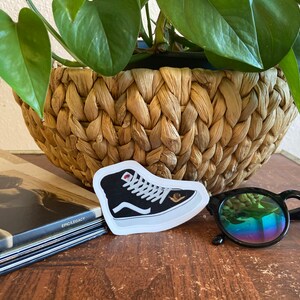May include: A black and white high-top sneaker sticker with a red accent. The sticker is placed on a wooden surface, next to a pair of sunglasses and a woven basket with a green plant.