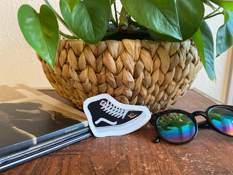 May include: A black and white high-top sneaker sticker with a red accent. The sticker is placed on a wooden surface, next to a pair of sunglasses and a woven basket with a green plant.