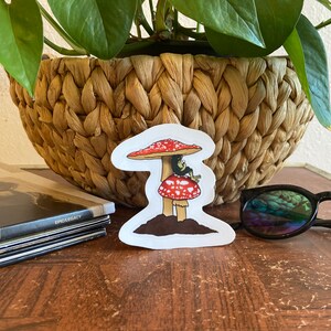 May include: A whimsical sticker featuring a red and white mushroom with a small creature perched on top. The sticker has a brown base and is set against a natural background. The sticker is a fun, decorative item.