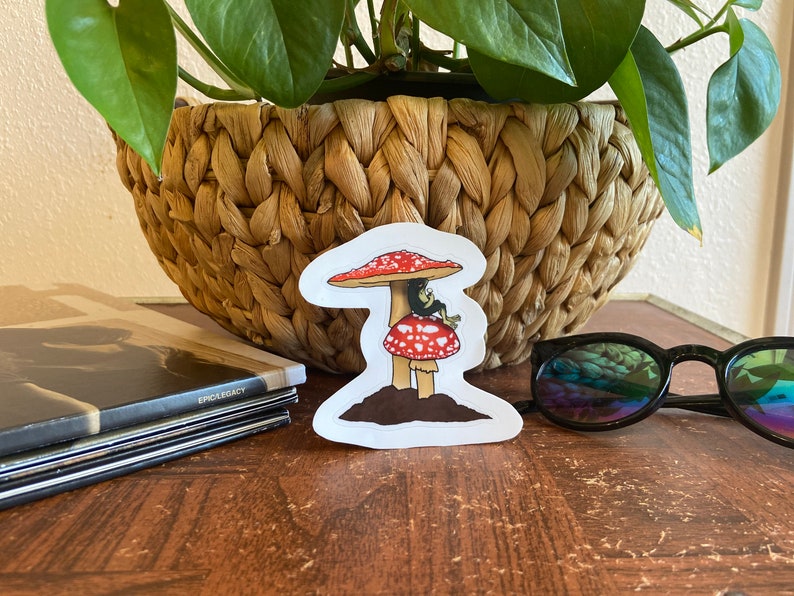 May include: A whimsical sticker featuring a red and white mushroom with a small creature perched on top. The sticker has a brown base and is set against a natural background. The sticker is a fun, decorative item.