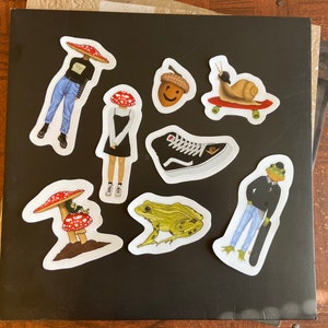 May include: A collection of whimsical stickers on a black surface. The stickers feature various designs, including a snail on a skateboard, a frog with a skateboard, a shoe, an acorn, and figures with mushroom caps. The stickers are colorful and detailed.