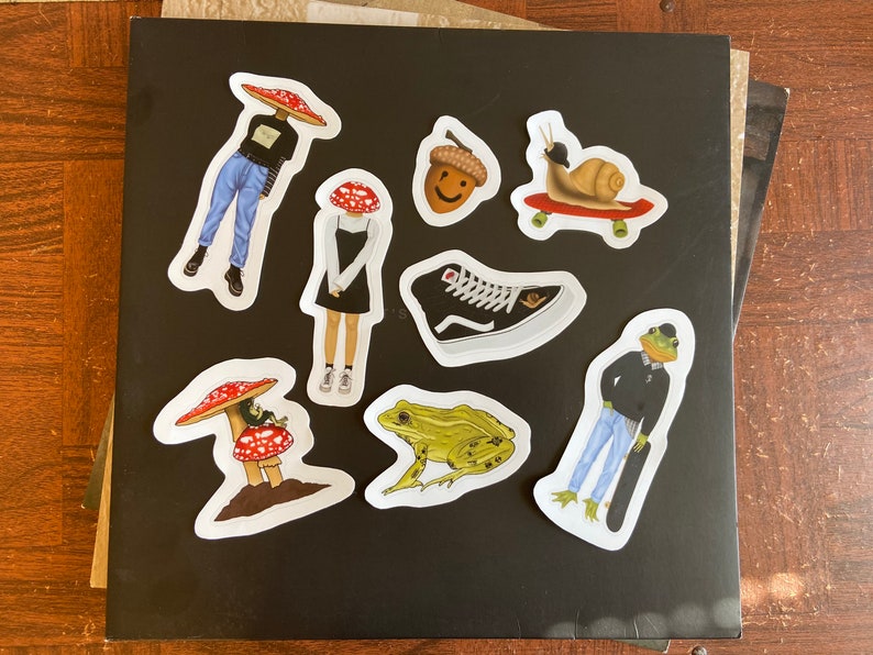 May include: A collection of whimsical stickers on a black surface. The stickers feature various designs, including a snail on a skateboard, a frog with a skateboard, a shoe, an acorn, and figures with mushroom caps. The stickers are colorful and detailed.