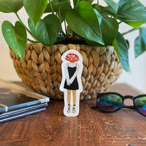 May include: A sticker featuring a cartoon figure with a mushroom head, wearing a black dress and sneakers. The sticker is placed on a wooden surface, near a woven basket with a green plant, a stack of books, and sunglasses.
