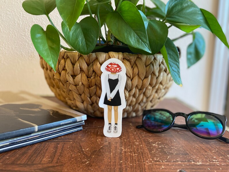 May include: A sticker featuring a cartoon figure with a mushroom head, wearing a black dress and sneakers. The sticker is placed on a wooden surface, near a woven basket with a green plant, a stack of books, and sunglasses.