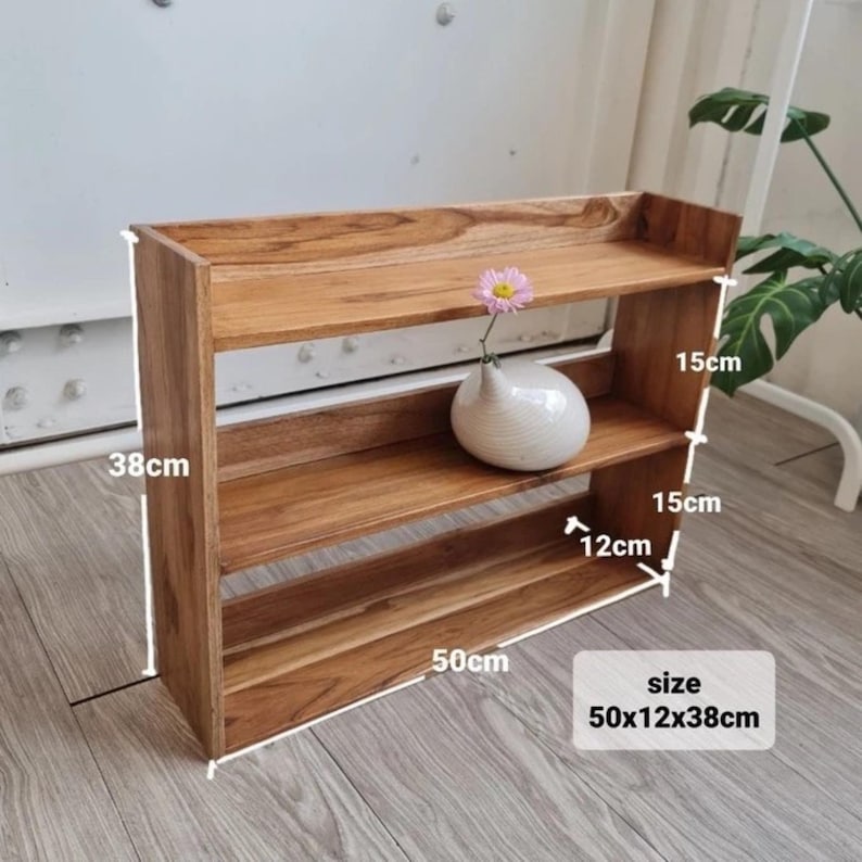 Op de afbeelding: Een houten plank met drie niveaus, 50 cm lang, 12 cm diep en 38 cm hoog. De natuurlijke houtnerf is zichtbaar. Een kleine witte vaas met een roze bloem staat op de bovenste plank.