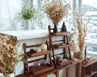 May include: Two-tiered wooden display shelves with a ladder design. The shelves hold decorative items such as glasses, candles, and a small Christmas tree. Dried plants and flowers in vases add to the natural, rustic aesthetic. The shelves are made of dark wood.