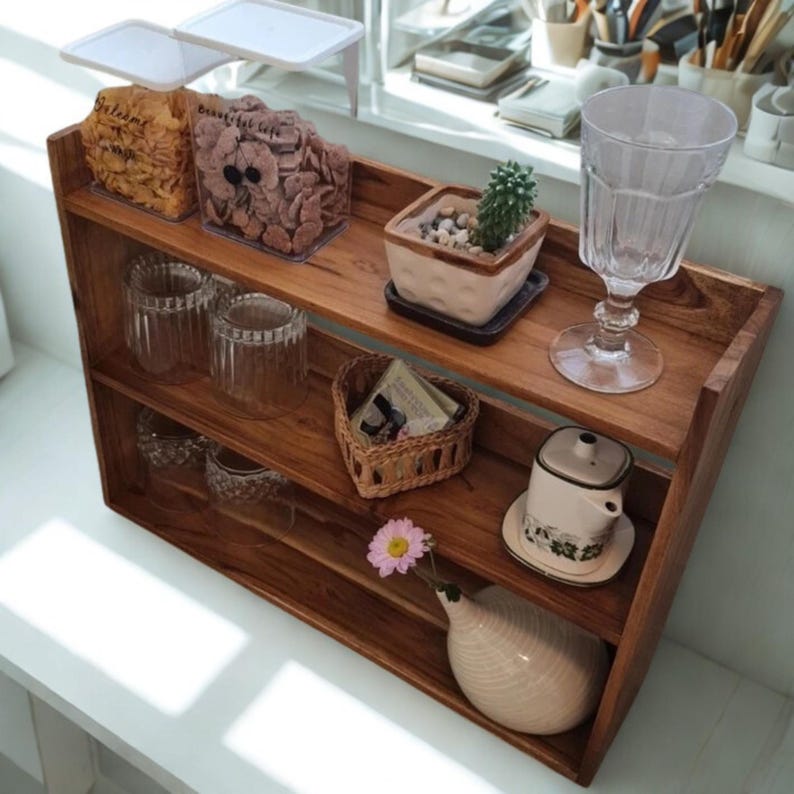 Op de afbeelding: Een houten plank met drie niveaus, met diverse items. De bovenste plank bevat doorzichtige bakjes met voedsel, een kleine cactus in een vierkante pot en een hoog glas. De onderste planken tonen glazen, een gevlochten mand en een kleine vaas met een bloem.
