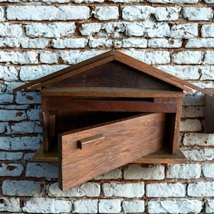 Può includere: Una cassetta delle lettere in legno con tetto a capanna e porta aperta, che rivela l'interno. La cassetta delle lettere è in legno marrone scuro ed è montata su un muro di mattoni bianchi.