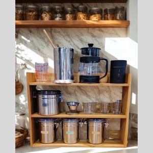 Op de afbeelding: Houten schap met koffie- en theeaccessoires. Inclusief een glazen cafeti&egrave;re, roestvrijstalen containers, heldere glazen kopjes en een zwarte mok. Potten met etenswaren staan op de bovenste plank.