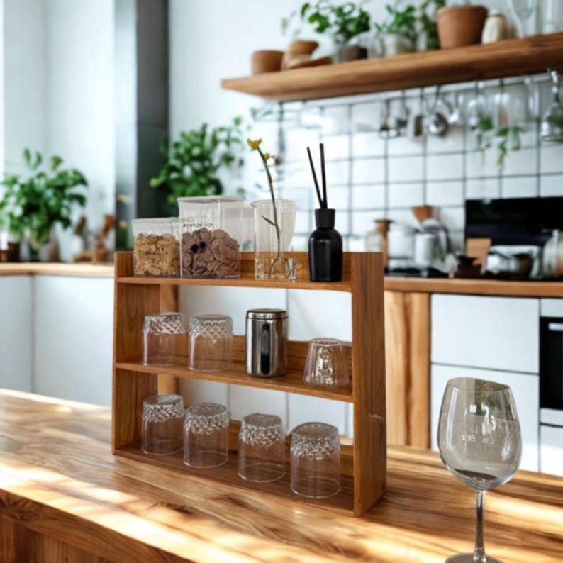 Op de afbeelding: Een houten keukenschap met twee niveaus, met heldere drinkglazen, een roestvrijstalen container en doorzichtige containers met voedsel. Een zwarte diffuserfles en een glazen vaas met een gele bloem staan ook op de plank. Een wijnglas staat op het houten aanrecht.