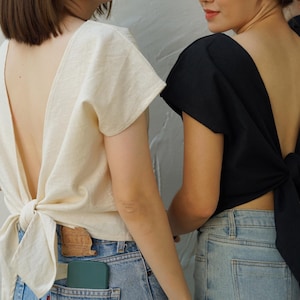 May include: Two women wearing tops with open backs that tie in the back. The woman on the left is wearing a cream-colored top and the woman on the right is wearing a black top. Both women are wearing blue jeans.