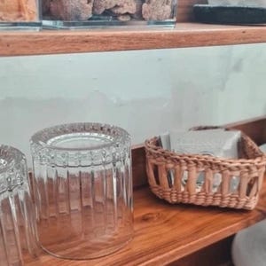 Op de afbeelding: Heldere, geribbelde drinkglazen en een klein geweven mandje op een houten plank. Het mandje bevat kleine, witte pakjes. De plank maakt deel uit van een grotere display, met glazen potten zichtbaar op de achtergrond.