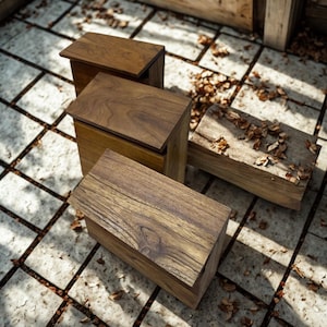 May include: Four wooden boxes with hinged lids, arranged on a tiled surface. The boxes are various sizes and have a natural wood grain finish. Some fallen leaves are scattered around the boxes, suggesting an outdoor setting.