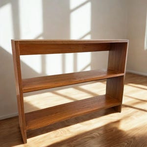 Op de afbeelding: Een houten boekenplank met drie niveaus en een warme, roodbruine afwerking. De planken zijn gelijkmatig verdeeld en worden ondersteund door zijpanelen. De boekenplank is vrijstaand en staat op een hardhouten vloer.